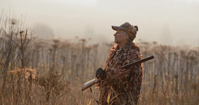 Portrait of hunter with shot gun standing in the field and looking around, foggy autumn morning, soft sun light