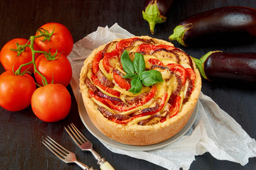 Traditional French quiche with vegetables and cheese on the black wooden background. Vegetarian tart decorated with fresh eggplants, tomatoes and basil leaves. Side view