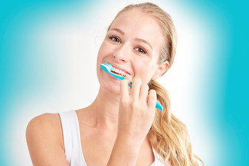 Studio shot of beautiful young woman brushing her teeth while looking at camera and smiling. Isolated on light blue background.