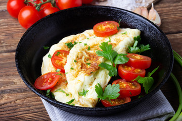 Omelet with cherry tomatoes and fresh green parsley in a black iron pan on a wooden background.