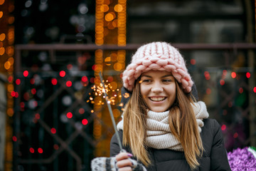 Positive blonde girl wearing winter outfit celebrating New Year at the street with sparklers. Space for text