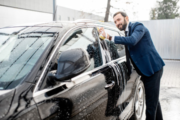Elegant businessman dressed in the suit washing his car with yellow sponge on a self service open...