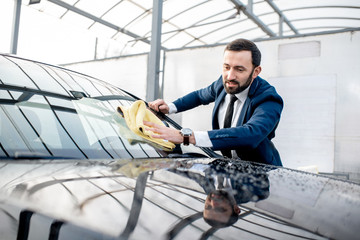 Smiling businessman dressed in a suit wiping windshield of his car with yellow microfiber on a self...