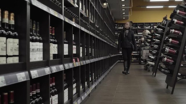 A young woman in the store is considering a shelf of wine to make a purchase.
