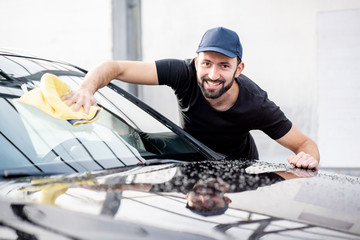 Handsome washer in t-shirt and cap wiping windshield with yellow microfiber at the open air car wash