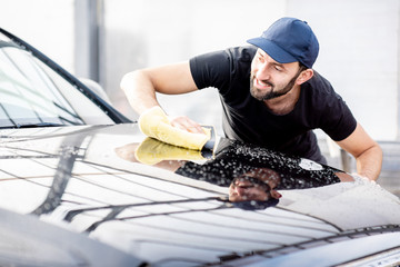 Professional washer in t-shirt and cap wiping a car hood with yellow microfiber at the open air car...