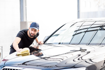 Professional washer in t-shirt and cap wiping a car hood with yellow microfiber at the open air car...