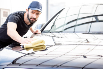 Professional washer in t-shirt and cap wiping a car hood with yellow microfiber at the open air car wash