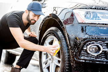Professional washer in black uniform and cap wiping with sponge car wheel during the washing...