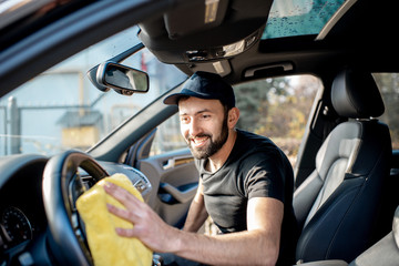 Handsome washer in black t-shirt and cap wiping helm in a luxury car interior with yellow microfiber