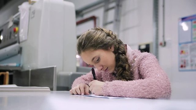 Teenager looking at camera and speaking. Cute Caucasian girl in pink sweater sitting at table and writing. Indoors.