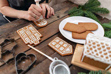 Icing of Christmas bakery. Woman decorating honey gingerbread cookies on wooden brown table....