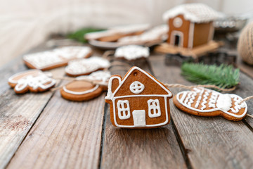 Cute little house in the foreground. A lot of ginger biscuits in different form on brown wooden table. Decorated with white sweet glaze. Christmas mood, winter morning. Fir branches