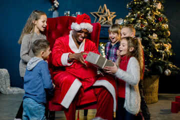 African Santa Claus takes gifts from a red bag for gifts and presents them to small, beautiful children.