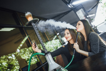 Two beautiful brunette friends are sitting on a summer terrace of a modern restaurant, chatting, doing selfie and smoking hookah, covered with blankets.