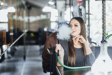 Fototapeta premium Young beautiful girl sits and smokes hookah on the summer terrace of a modern cafe.