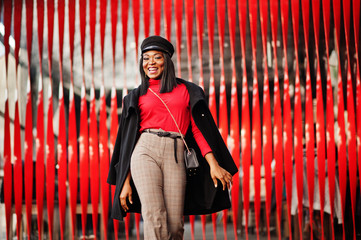 African american fashion girl in coat and newsboy cap posed at street against red background.