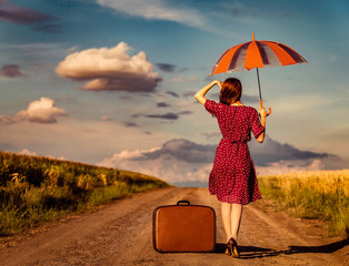 Redhead girl in red dress with hat and suitcase holding umbrella and walking down on rural road...