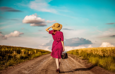Redhead girl in red dress with hat and suitcase walking down on rural road near wheat field and cloudscape on background.