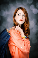 Beautiful surprised caucasian redhead adult girl in coral shirt and shopping bags on gray backgroud.