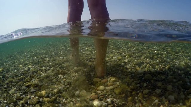 Sexy fashion model in bikini walking in shallow sea water toward camera on beach sith stones, gopro dome shot