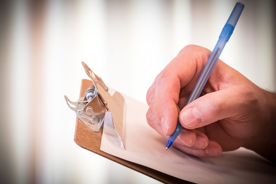 Man's hand puts pen to paper on a clipboard, to sign a form, make a list, or fill out information.
