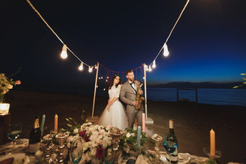 Wedding banquet on the ocean shore at night. The groom is playing the saxophone.