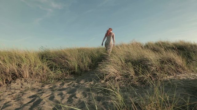 A woman walks on a narrow footpath among beach dunes by summer.