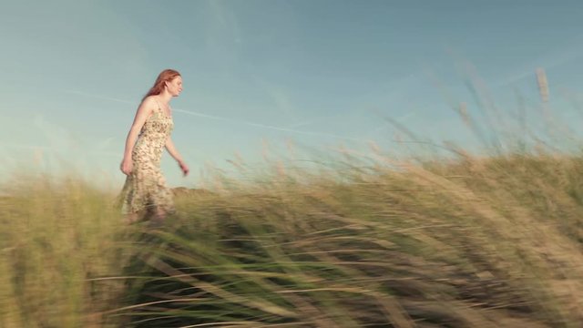 Tracking footage of a young woman walking among dune grass on a beach by summer.