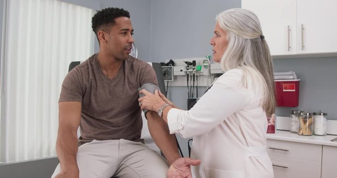 Senior female doctor checking young black males blood pressure with monitor and stethoscope. Mature caucasian doctor checking blood pressure of young african-american patient