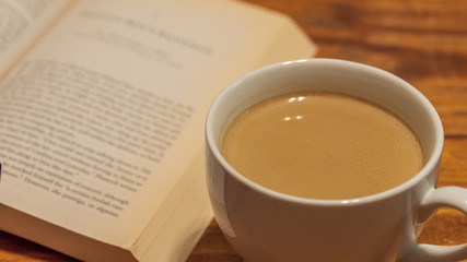 A white ceramic cup of coffee with cream next to a book, sitting atop a wooden coffee table