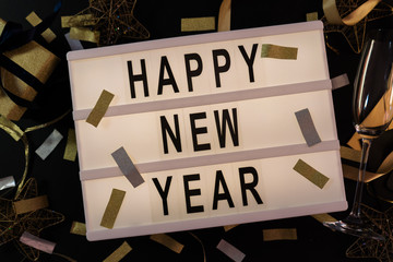 Happy New Year sign on a lightbox with golden confetti decoration on black background. New Years Eve party. Flat lay. Top view. 2019