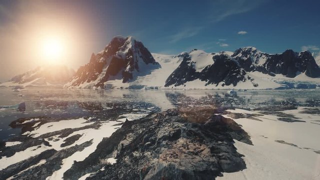 Sunset over Antarctica Mountains, Ocean. Lemaire Channel Aerial Flight. Drone Overview Shot Of Mighty Mounts, Snow, Ice Covered Land. Bright Sun Over Ice Cold Ocean. Winter Landscape. 4k Footage.
