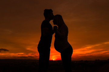 Silhouettes of husband kissing his pregnant wife into forehead and enjoy spending time together outdoor.