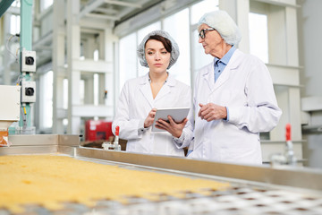 Waist up portrait  of  two workers doing  production quality inspection in food factory standing by conveyor belt and using digital tablet, copy space