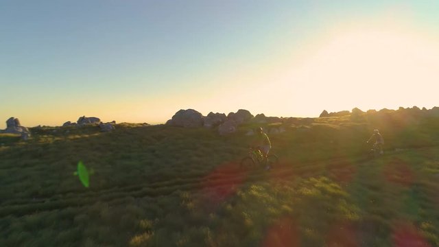 AERIAL, LENS FLARE: Young couple mountain biking along the scenic path on a sunny morning. Male and female tourists pedal their bicycles up a grassy hill on a beautiful evening. Fun outdoor activity.