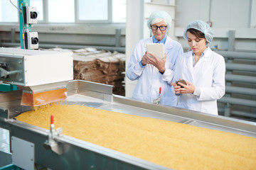 Waist up portrait of two female factory workers standing by macaroni conveyor belt during quality...