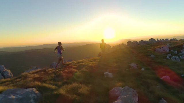 DRONE, SUN FLARE: Flying behind sporty tourist couple running along mountain trail at sunset. Golden morning sunbeams shine on athletic young man and woman training for a difficult fell running race.