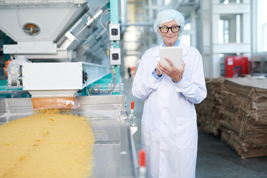 Portrait of senior woman working at food factory standing by conveyor belt and using digital tablet, copy space
