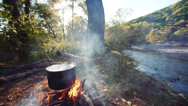 Beautiful view of peaceful camping under big tree with burning campfire and pot with boiling soup above it. Fast blue stream and october forest are on background. Far East of Russia. Nice sunny day