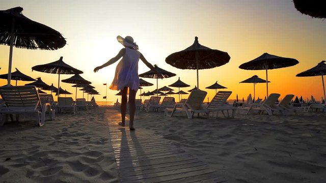 Woman in white dress and hat walk toward camera on beach in the seaside resort with sun loungers and wooden sheds against sunset, cinematic