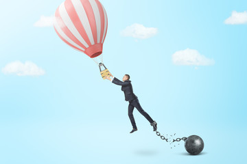 A businessman breaks away from a chain and iron ball to catch a small hot air balloon.