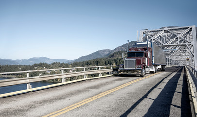 Big rig burgundy semi truck with shiny refrigerator semi trailer driving on the truss bridge in Columbia Gorge