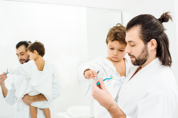 Toddler son looking at toothbrush with toothpaste and dad holding him in white bathroom