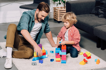 man with little son playing with colorful plastic blocks on floor at home