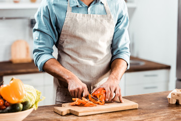 mid section of young man in apron cutting fresh pepper in kitchen