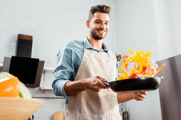 low angle view of smiling young man in apron holding frying pan with vegetables