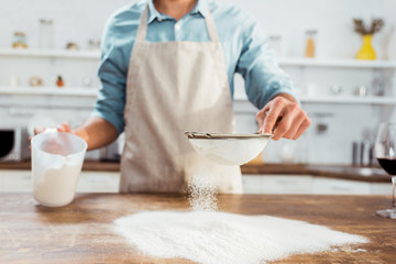 partial view of young man in apron sifting flour on kitchen table