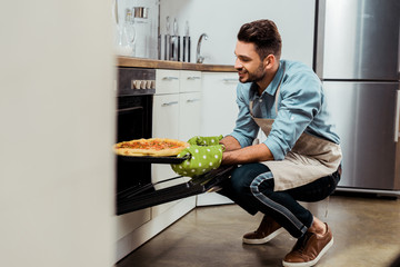 smiling young man in apron taking out baking tray with pizza from oven