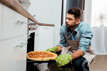 young man in apron and potholders taking out baking tray with pizza from oven